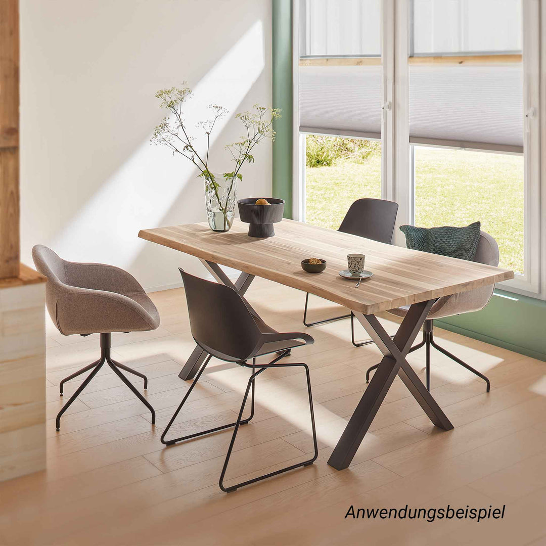 Ein massiver Esstisch mit heller Holzplatte und einem X-förmigen Untergestell aus schwarzem Stahl, der in einem lichtdurchfluteten Raum steht. Das Arrangement wird durch vier graue Schalenstühle und eine schlichte, moderne Tischdekoration abgerundet.