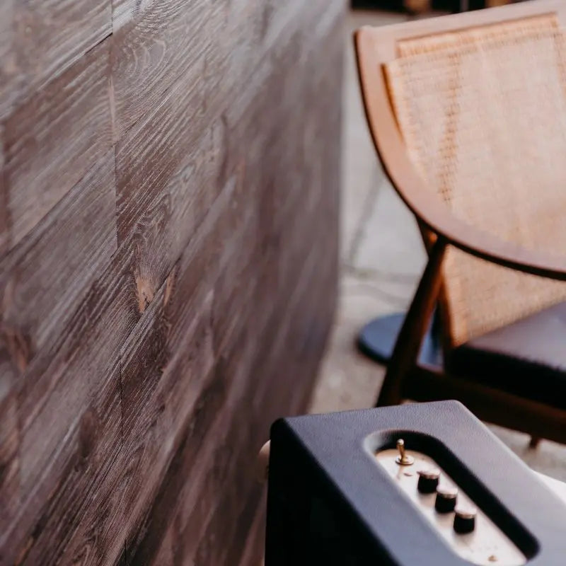 Nahaufnahme einer dunkelbraunen Holzwand neben einem Rattanstuhl und einem Lautsprecher. Die sichtbare Struktur des Holzes unterstreicht die warme Raumatmosphäre.