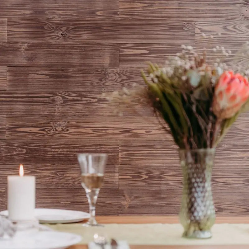 Wandverkleidung mit dunklen Holzpaneelen „Ur“, davor ein gedeckter Tisch mit Blumen, Gläsern und einer Kerze