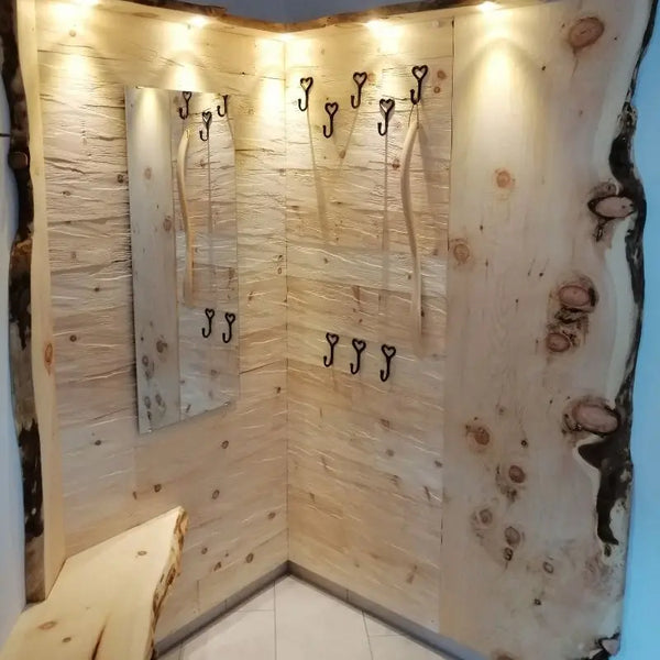 Eine Garderobe ist mit massiven, hellen Zirbenholzbrettern verkleidet, die deutliche Astlöcher und eine natürliche Baumkante aufweisen. Integrierte Deckenspots, ein Spiegel und herzförmige Kleiderhaken ergänzen die rustikale Flurgarderobe.
