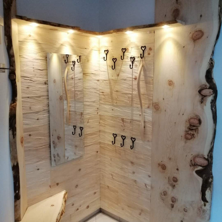 Eine individuell gefertigte Garderobenecke, die komplett mit hellen Holzpaneelen ausgekleidet ist und über geschwungene Echtholzkanten verfügt. Schwarze Kleiderhaken in Herzform, ein integrierter Spiegel und dezente Spotlichter betonen die natürliche und hochwertige Verarbeitung des Materials.