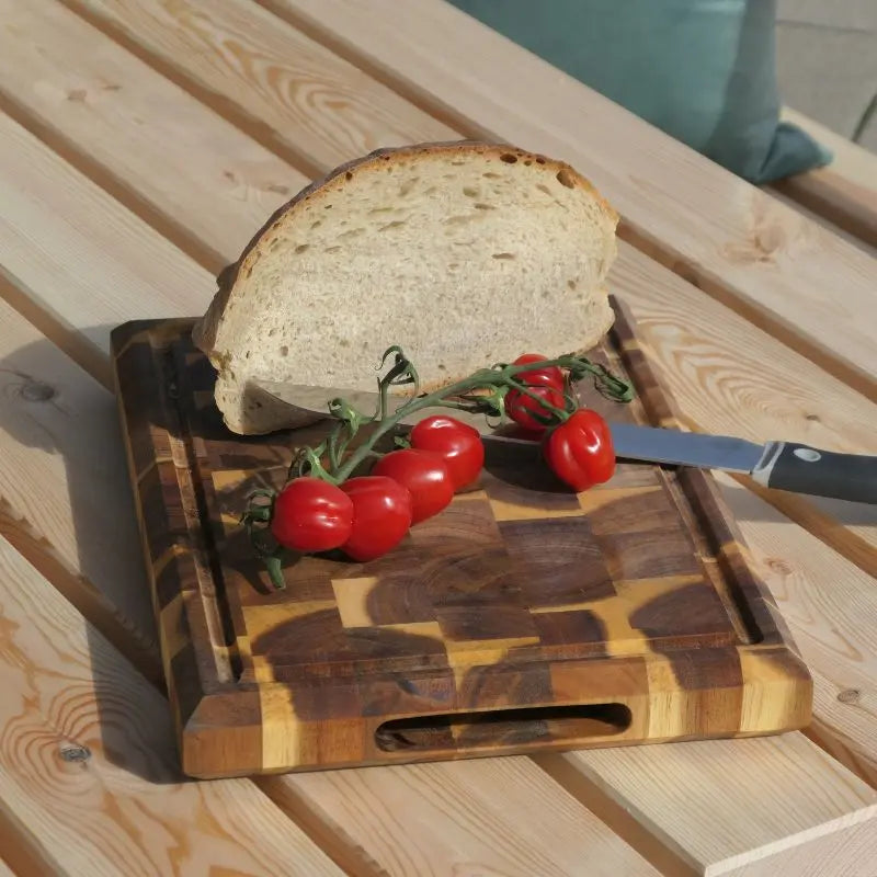 Ein robustes Schneidebrett mit deutlich sichtbarer, schachbrettartiger Stirnholz-Maserung. Auf dem Brett liegen eine dicke Scheibe Brot, Rispentomaten und ein Messer.