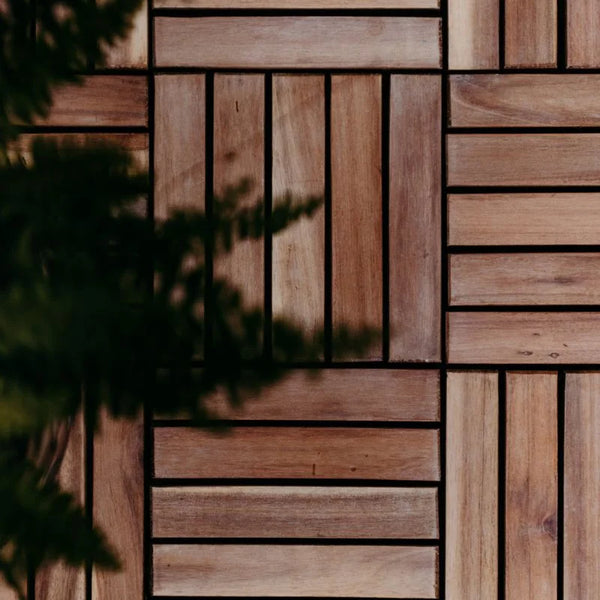 Nahaufnahme von quadratischen Akazienholz Terrassenfliesen aus braunem Holz, die in einem abwechselnden Schachbrettmuster verlegt sind. Dunkelgrüne Pflanzenblätter ragen unscharf in den linken Bildrand.