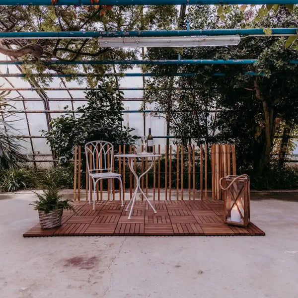 Ein mit Klickfliesen aus Thermo-Esche ausgelegter Boden, auf dem ein kleiner Metalltisch und ein Metallstuhl sowie eine Pflanze und ein Windlicht stehen.