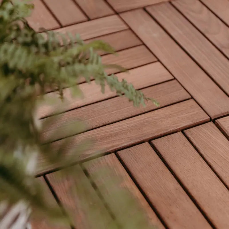 Detailansicht von dunkel-schokobraunen Terrassenfliesen, die passgenau im Schachbrettmuster verlegt sind. Ein grüner Farnwedel ragt unscharf in den linken Bildrand.