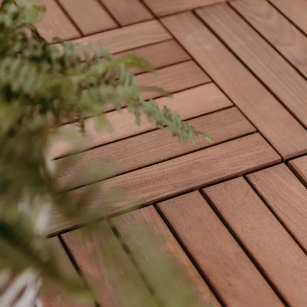 Detailansicht von dunkel-schokobraunen Terrassenfliesen, die passgenau im Schachbrettmuster verlegt sind. Ein grüner Farnwedel ragt unscharf in den linken Bildrand.