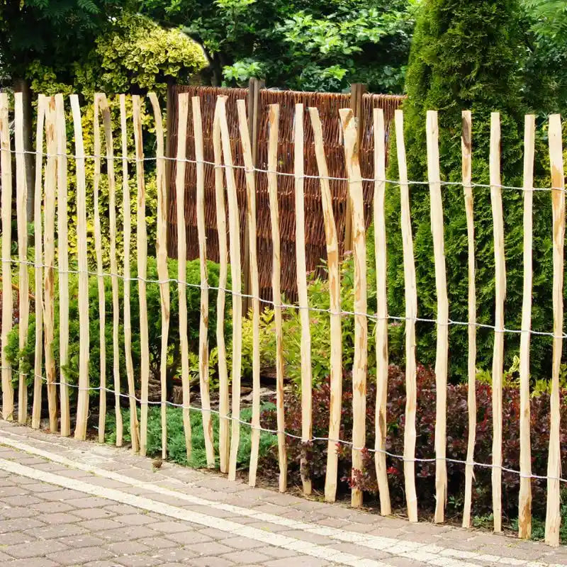 Ein robuster Staketenzaun aus naturbelassenem Haselnussholz grenzt einen gepflasterten Gehweg von einem dicht bepflanzten Gartenbereich ab. Die ungleichmäßigen, witterungsbeständigen Holzlatten unterstreichen die natürliche Optik im Außenbereich.