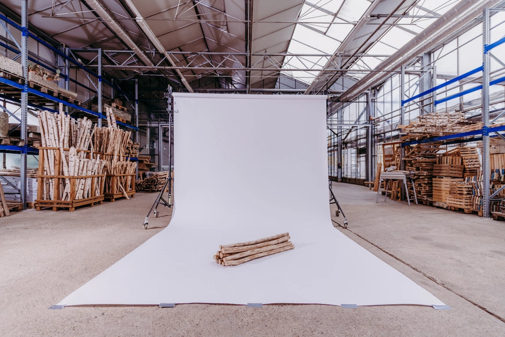 Ein professioneller weißer Fotohintergrund ist mitten in einer großen Lagerhalle aufgebaut, auf dem ein Stapel kleiner Holzstämme für ein Produktfoto bereitliegt. Im Hintergrund sind Regale mit zahlreichen Birkenstämmen und Holzvorräten der Werkstatt zu sehen.