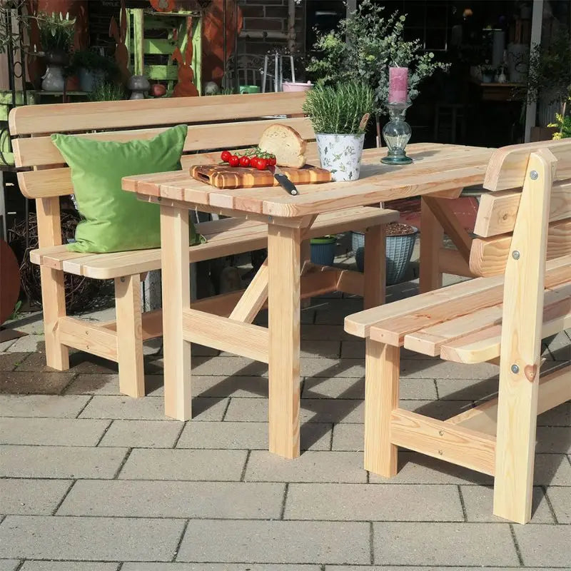 Eine robuste Gartensitzgruppe aus hellem, naturbelassenem Holz steht auf einer gepflasterten Terrasse. Auf dem Tisch liegen ein Laib Brot und Rispentomaten auf einem Schneidebrett bereit.