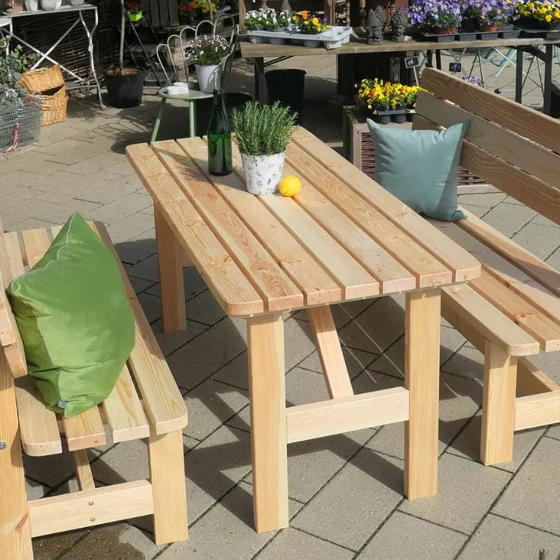 Eine rustikale Gartengarnitur aus hellem Massivholz mit einem großen Tisch und zwei passenden Sitzbänken, die mit grünen Kissen dekoriert sind. Das robuste Möbel-Set steht im Freien auf einer gepflasterten Fläche und bietet viel Platz für gesellige Runden.