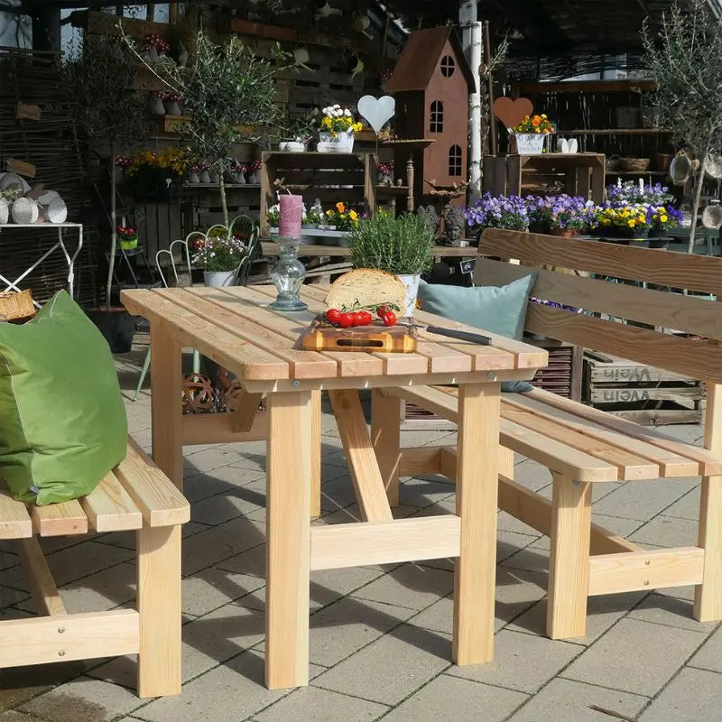 Eine gemütlich arrangierte Sitzgruppe aus hellem Kiefernholz mit grünen Kissen lädt auf einer Terrasse zum Verweilen ein. Im Hintergrund befindet sich ein liebevoll dekorierter Gartenbereich mit zahlreichen Blumen und Topfpflanzen.