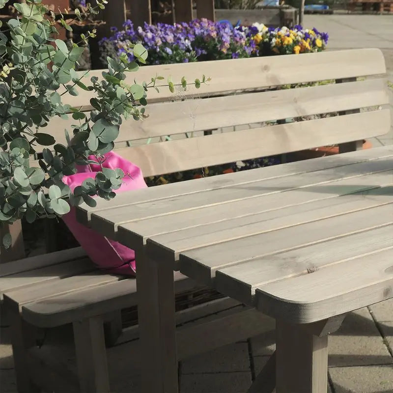 Detailansicht einer stabilen, grau lasierten Gartenbank und eines passenden Tisches aus Kiefernholz im sonnigen Außenbereich. Im linken Vordergrund ragt eine dekorative Grünpflanze ins Bild.