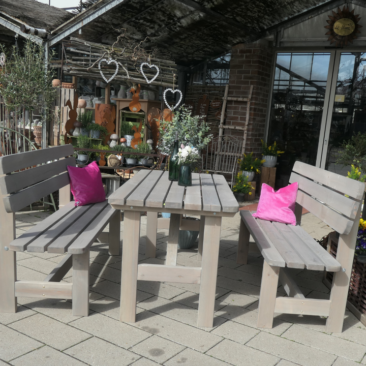 Eine Gartengarnitur aus grau lasiertem Kiefernholz mit Tisch und zwei Bänken auf einer hell gepflasterten Terrasse. Zwei leuchtend pinke Kissen setzen auf den Bänken farbliche Akzente.