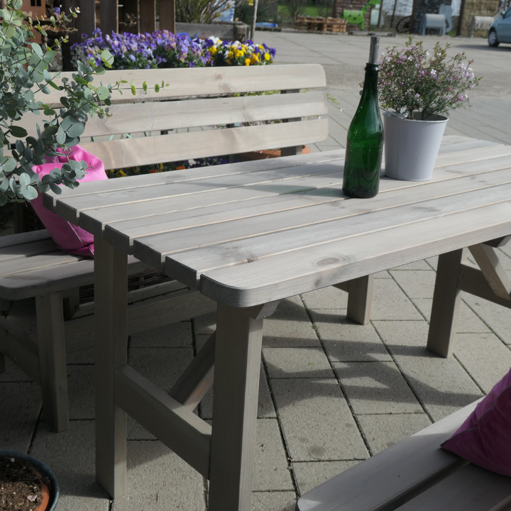 Eine rustikale Garten-Sitzgruppe aus grau lasiertem Holz, bestehend aus einem Latten-Tisch und passenden Bänken auf einer gepflasterten Terrasse. Auf dem Tisch stehen eine grüne Weinflasche und ein kleiner Pflanztopf, während pinke Kissen für farbige Akzente auf den Bänken sorgen.