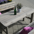 Eine rustikale Garten-Sitzgruppe aus grau lasiertem Holz, bestehend aus einem Latten-Tisch und passenden Bänken auf einer gepflasterten Terrasse. Auf dem Tisch stehen eine grüne Weinflasche und ein kleiner Pflanztopf, während pinke Kissen für farbige Akzente auf den Bänken sorgen.
