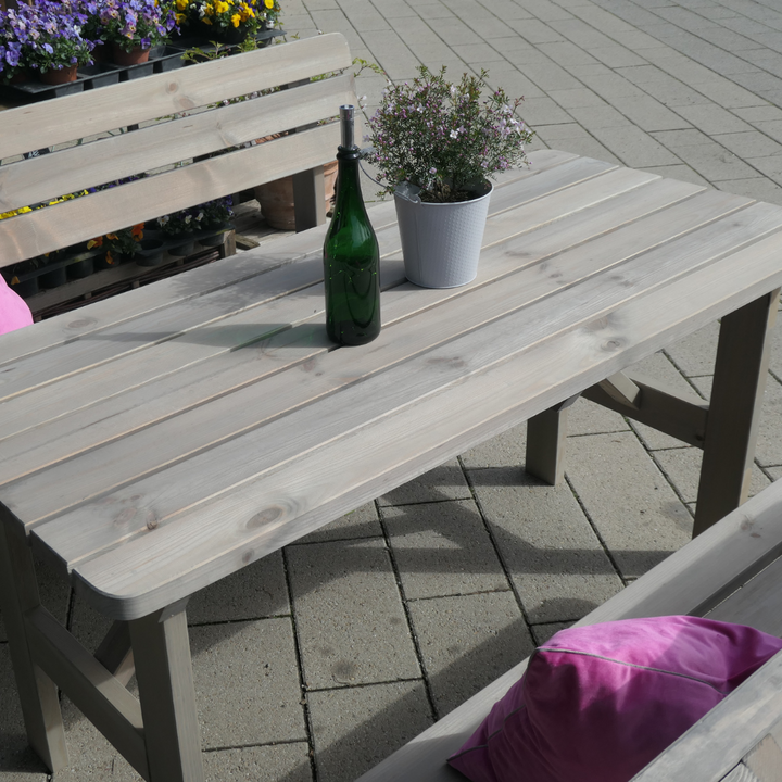 Eine rustikale Garten-Sitzgruppe aus grau lasiertem Holz, bestehend aus einem Latten-Tisch und passenden Bänken auf einer gepflasterten Terrasse. Auf dem Tisch stehen eine grüne Weinflasche und ein kleiner Pflanztopf, während pinke Kissen für farbige Akzente auf den Bänken sorgen.