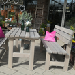 Eine Gartengarnitur aus grau lasiertem Kiefernholz mit Tisch und zwei Bänken auf einer hell gepflasterten Terrasse. Zwei leuchtend pinke Kissen setzen auf den Bänken farbliche Akzente.