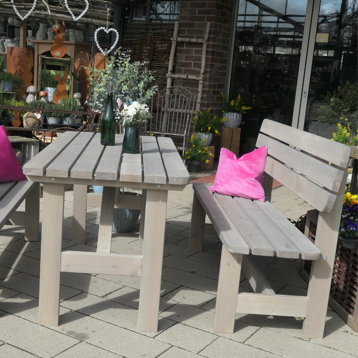 Eine Gartengarnitur aus grau lasiertem Kiefernholz mit Tisch und zwei Bänken auf einer hell gepflasterten Terrasse. Zwei leuchtend pinke Kissen setzen auf den Bänken farbliche Akzente.