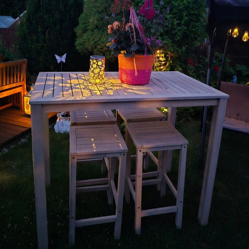 Ein Hochtischset aus Akazienholz mit einer brennenden Kerze und einem Blumengesteck in der Dämmerung.