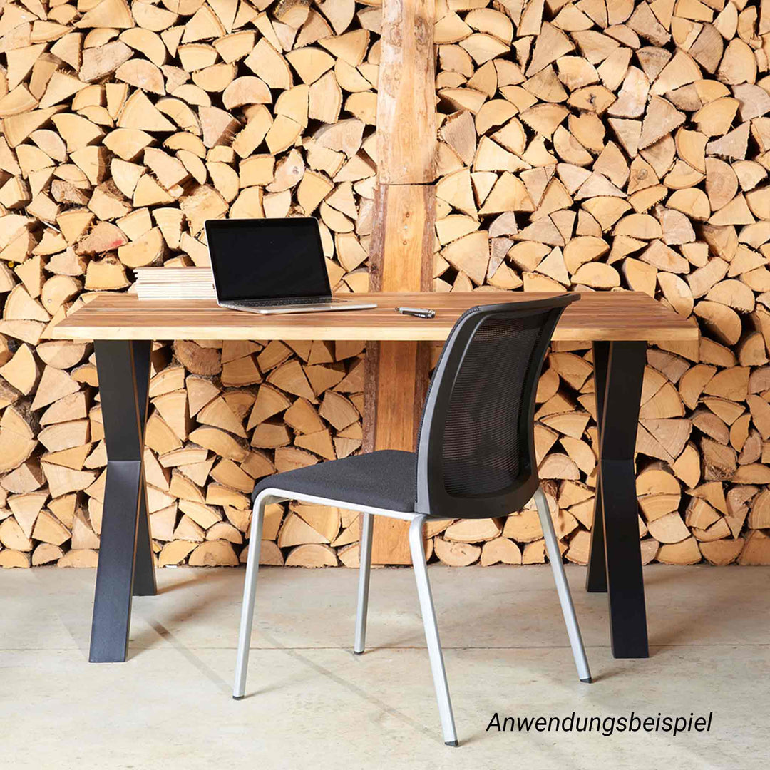Ein moderner Schreibtisch mit massiver Holzplatte und schwarzem Metallgestell bildet einen stilvollen Kontrast zu einer raumhohen Wand aus gestapeltem Brennholz. Ein schwarzer Bürostuhl und ein aufgeklappter Laptop auf dem Tisch ergänzen diesen rustikal-modernen Arbeitsplatz.