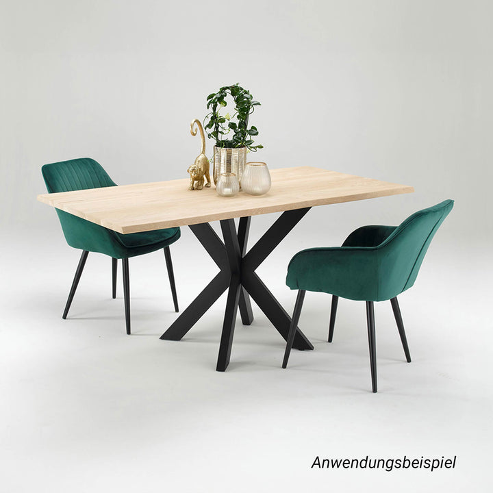 Ein Esstisch mit massiver, heller Holzplatte und mattschwarzem Spider-Gestell aus Metall, umgeben von zwei dunkelgrünen Polsterstühlen mit Armlehnen. Auf dem Tisch arrangierte goldene Deko-Objekte und eine Pflanze setzen elegante Akzente zum modernen Industrial-Look des Untergestells.
