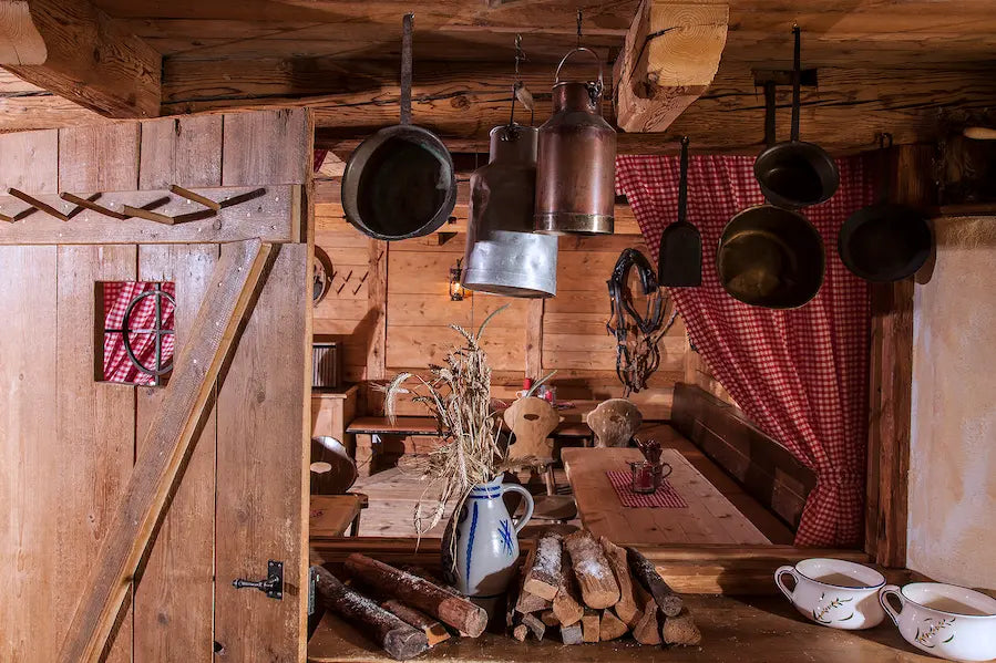Ein Blick durch eine geöffnete Holztür in eine rustikale Stube, in der antike Kupferkessel und Pfannen von der Decke hängen. Im Vordergrund ergänzen Brennholz, ein Steingutkrug und weißes Geschirr das historische, ländliche Dekor.