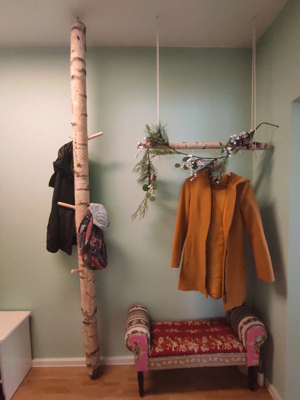 Eine kreative Garderobe aus einem vertikalen Birkenstamm mit Holzhaken und einer horizontal hängenden Birke als Kleiderstange. Die naturverbundene Gestaltung vor einer mintgrünen Wand wird durch eine kleine, floral gemusterte Sitzbank im Vintage-Stil ergänzt.