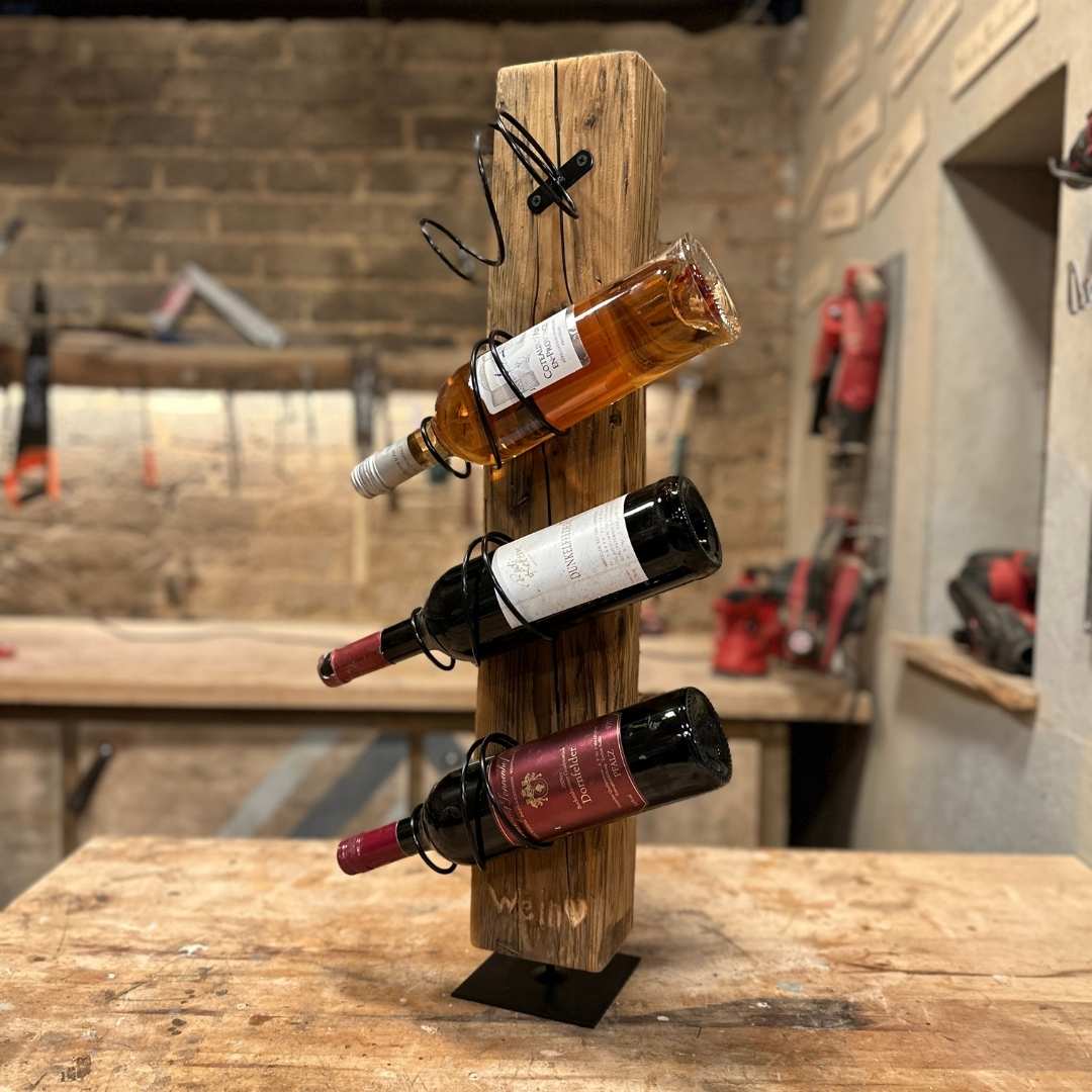 Ein handgefertigtes Weinregal aus einem senkrechten Altholzbalken, in dessen geschwungenen Metallhalterungen drei Weinflaschen lagern. Das Regal steht auf einem quadratischen schwarzen Metallfuß inmitten einer rustikalen Werkstattumgebung.