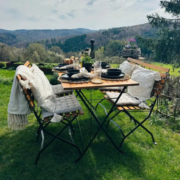 Ein einladend gedeckter Holztisch mit passenden Klappstühlen steht in einem grünen Garten vor einer weiten, bewaldeten Hügellandschaft. Helle Sitzkissen und schwarzes Geschirr auf Naturfaser-Platzsets unterstreichen das gemütliche Ambiente im Freien.