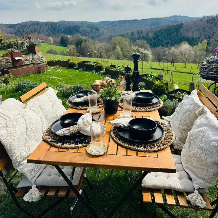 Eine detaillierte Ansicht eines gedeckten Gartentisches aus Holz mit schwarzen Tellern, Trinkgläsern und Leinenservietten auf geflochtenen Untersetzern. Im unscharfen Hintergrund erstreckt sich eine grüne Landschaft unter einem leicht bewölkten Himmel.