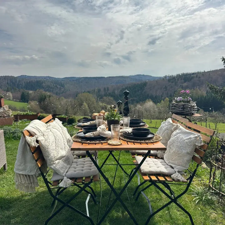 Eine rustikale Holz-Sitzgruppe für vier Personen, angerichtet für ein Essen im Freien, bietet einen weiten Blick über ein malerisches Tal. Die Kombination aus warmem Holz, hellen Textilien und der weitläufigen Natur strahlt Ruhe und Entspannung aus.