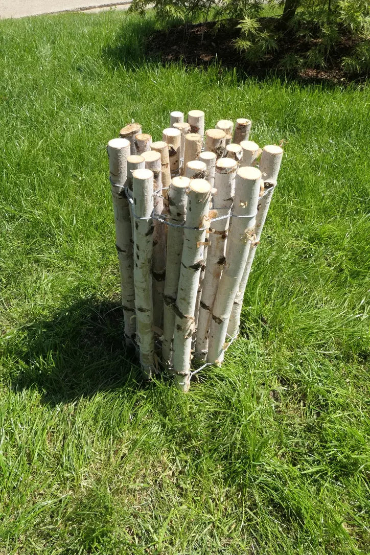 Ein vertikales Bündel aus mehreren hellen Birkenstämmen ist mit Draht fixiert und steht als dekoratives Element auf einer grünen Rasenfläche. Im Hintergrund ist ein kleiner Baum sowie ein sonniges Gartenambiente zu erkennen.
