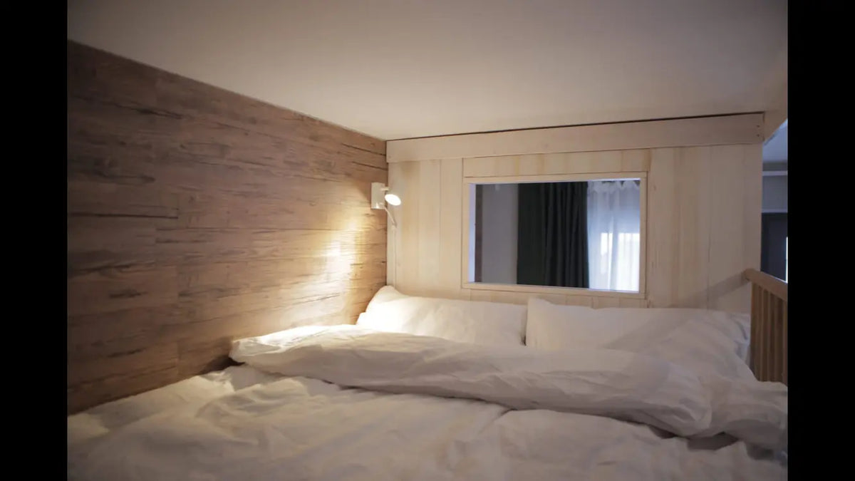 Ein gemütliches Bett mit weißer Bettwäsche, das direkt an eine Wand mit warmer Holzverkleidung grenzt. Eine kleine Wandleuchte und ein integriertes Fenster in der hellen Nebenwand schaffen eine behagliche und moderne Schlafatmosphäre.