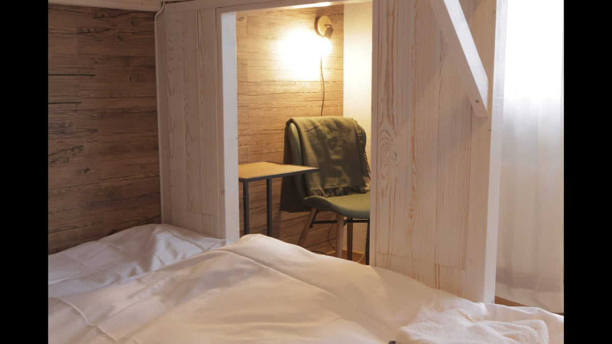 Der Blick aus einem gemütlichen Bett mit weißer Wäsche auf einen kleinen, integrierten Arbeitsplatz in einer Nische. Ein schlichter Tisch und ein grüner Sessel stehen vor einer Wand mit warmer Holzverkleidung, die durch eine dezente Wandleuchte stimmungsvoll erhellt wird.