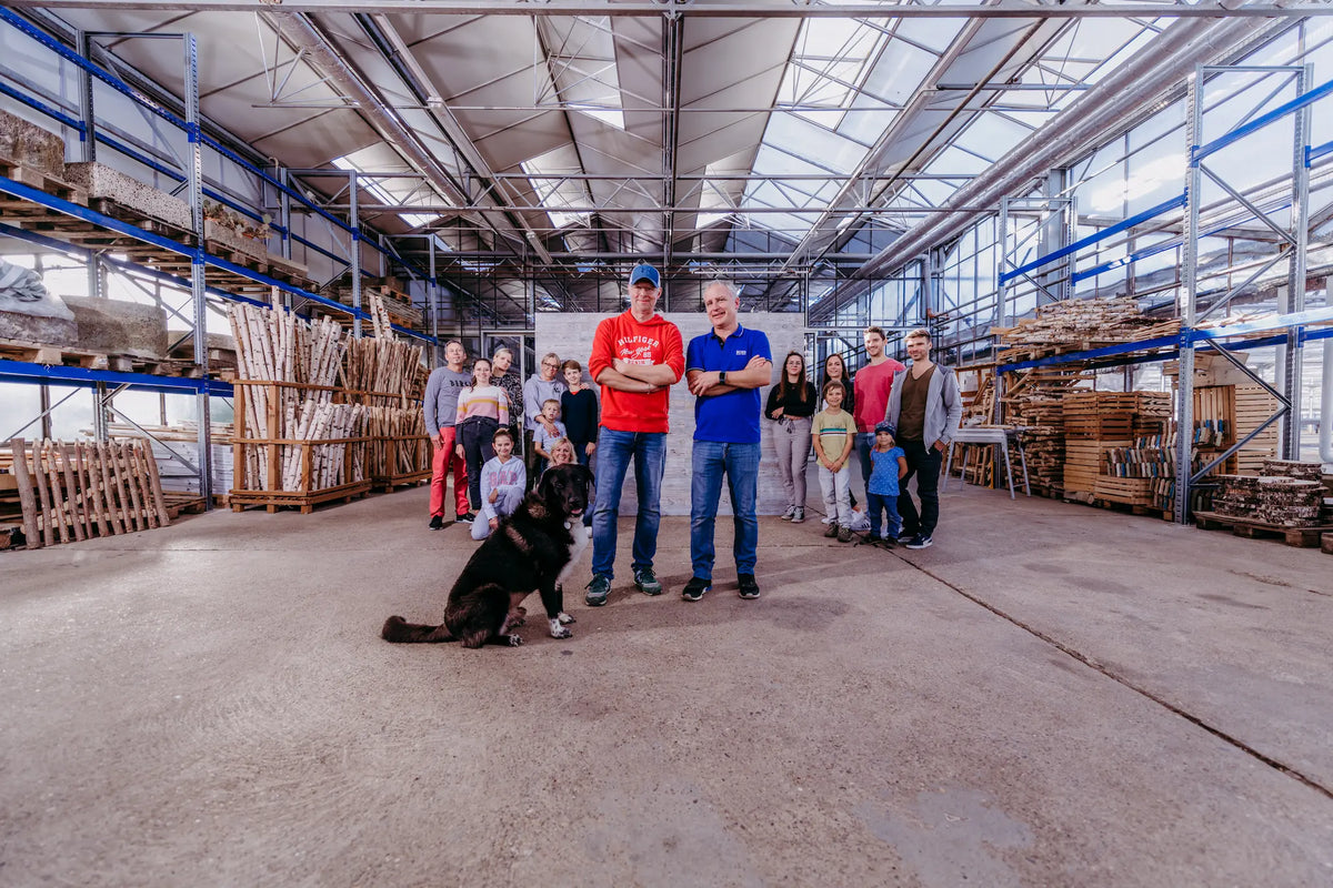 Das Team von iWerk24 posiert gemeinsam in einer großen Produktionshalle, in der im Hintergrund zahlreiche Regale mit Birkenstämmen und Holzvorräten gelagert sind. Ein großer schwarzer Hund sitzt entspannt im Vordergrund vor der Gruppe.