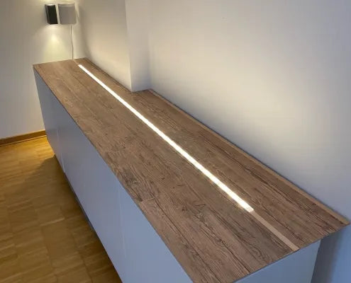 Ein langes, weißes Sideboard, das mit Holzpaneelen verkleidet wurde. Ein mittig eingelassener, leuchtender LED-Streifen zieht sich über die gesamte Länge der Holzoberfläche und setzt einen modernen Lichtakzent.