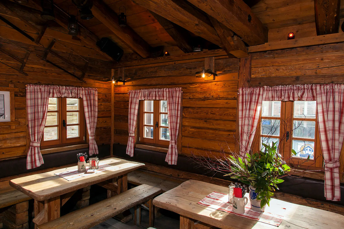 Der Innenraum einer gemütlichen Almhütte mit massiven Holzwänden, schweren Deckenbalken und Holztischen. Rot-weiß karierte Vorhänge an den kleinen Fenstern und warmes Licht erzeugen ein einladendes, traditionelles Ambiente.