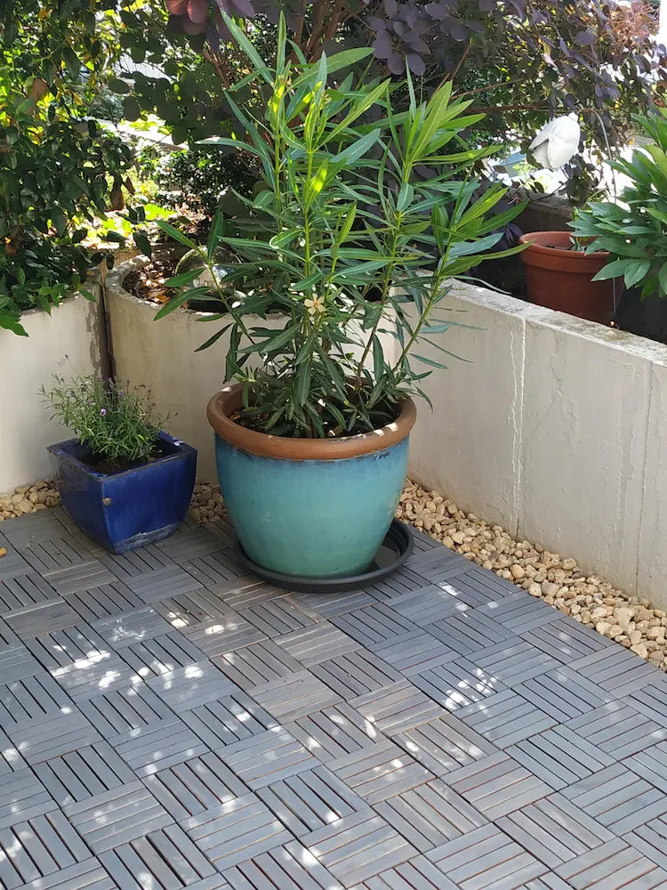Eine sonnige Terrasse, die mit grauen Holz-Klickfliesen in einem dekorativen Schachbrettmuster ausgelegt ist. Auf dem Boden stehen zwei große Pflanztöpfe, die den Außenbereich harmonisch begrünen.
