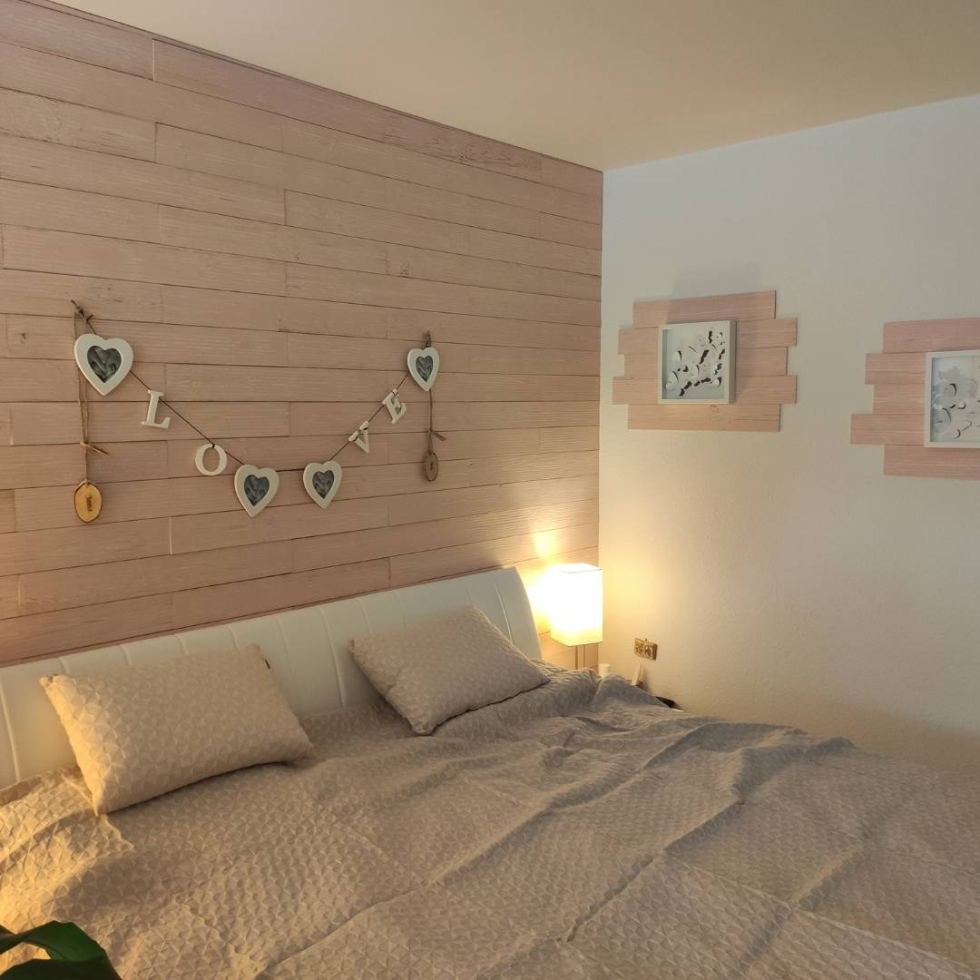 Ein modern eingerichtetes Schlafzimmer, bei dem die Wand hinter dem Bett mit horizontalen, hellrosa lasierten Holzpaneelen verkleidet ist. Zur Dekoration hängen eine "LOVE"-Girlande sowie farblich passende Holzelemente mit Bilderrahmen an der Wand.