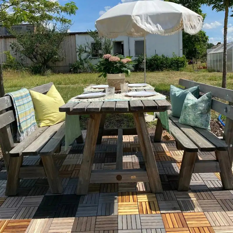 Ein rustikaler Holztisch mit Sitzbänken und einem weißen Sonnenschirm steht stabil auf einer Terrasse aus gemischten Holz-Klickfliesen. Die Szenerie im grünen Garten verdeutlicht die hohe Tragfähigkeit des Bodenbelags für schwere Außenmöbel.