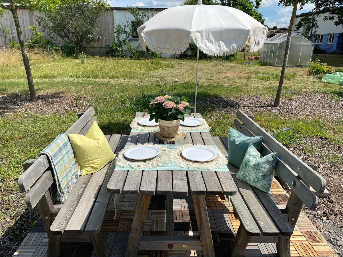 Eine einladende Outdoor-Sitzgruppe aus Holz steht auf einer Terrasse, die mit abwechselnd gemusterten Holz-Klickfliesen ausgelegt ist. Der für vier Personen gedeckte Gartentisch mit bunten Sitzkissen und einer Hortensie veranschaulicht eine sommerliche Wohlfühlatmosphäre im Freien.