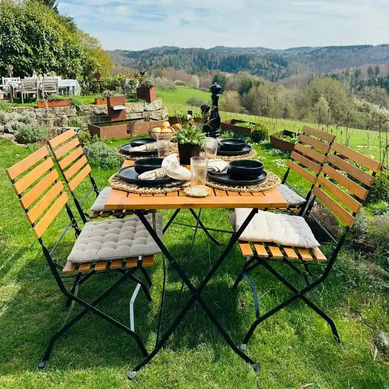 Ein sommerlich gedeckter Gartentisch mit vier passenden Klappstühlen steht auf einer grünen Rasenfläche vor einem weiten Landschaftspanorama.