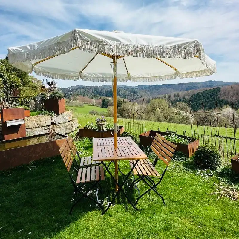 Ein sommerlich gedeckter Gartentisch mit vier passenden Klappstühlen steht auf einer grünen Rasenfläche vor einem weiten Landschaftspanorama.