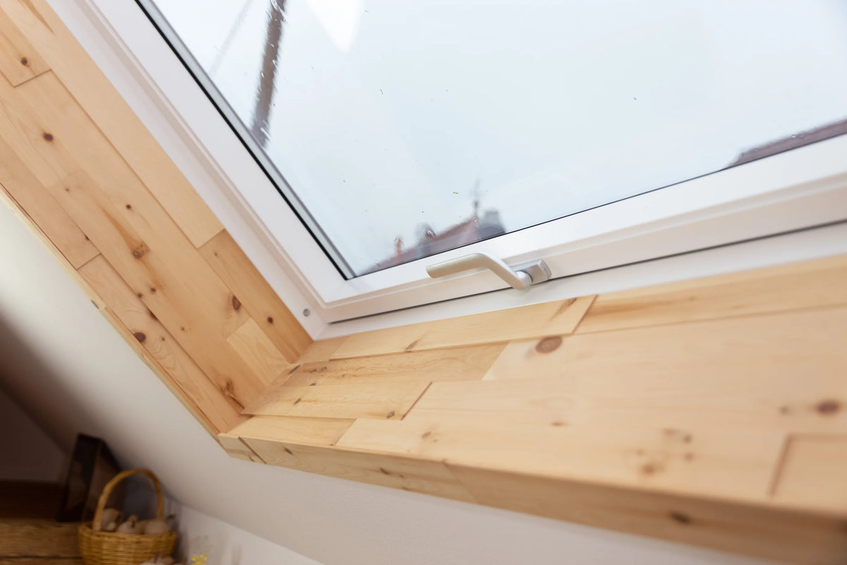 Ein weißes Dachfenster, dessen Innenlaibung komplett mit hellen Holzpaneelen ausgekleidet wurde. Die präzise verarbeiteten Paneele verleihen dem Fensterausschnitt eine natürliche Wärme und rahmen den Ausblick auf die gegenüberliegenden Dächer ein.