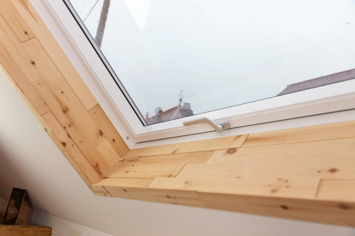 Ein weißes Dachfenster, dessen Innenlaibung komplett mit hellen Holzpaneelen ausgekleidet wurde. Die präzise verarbeiteten Paneele verleihen dem Fensterausschnitt eine natürliche Wärme und rahmen den Ausblick auf die gegenüberliegenden Dächer ein.