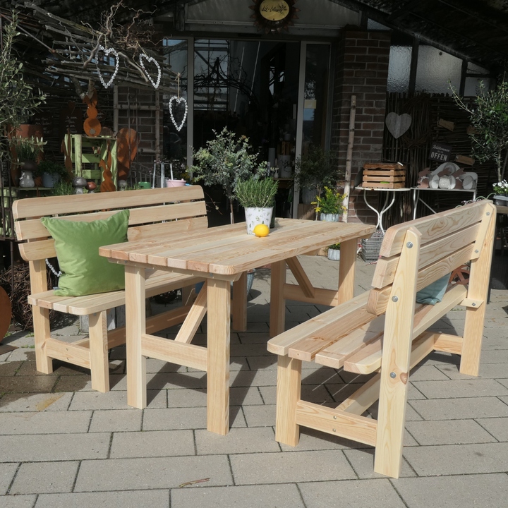 Eine rustikale Gartengarnitur aus hellem Massivholz mit einem großen Tisch und zwei passenden Sitzbänken, die mit grünen Kissen dekoriert sind. Das robuste Möbel-Set steht im Freien auf einer gepflasterten Fläche und bietet Platz für gesellige Runden.