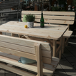 Eine rustikale Gartengarnitur aus hellem Massivholz mit einem großen Tisch und zwei passenden Sitzbänken, die mit grünen Kissen dekoriert sind. Das robuste Möbel-Set steht im Freien auf einer gepflasterten Fläche und bietet viel Platz für gesellige Runden.