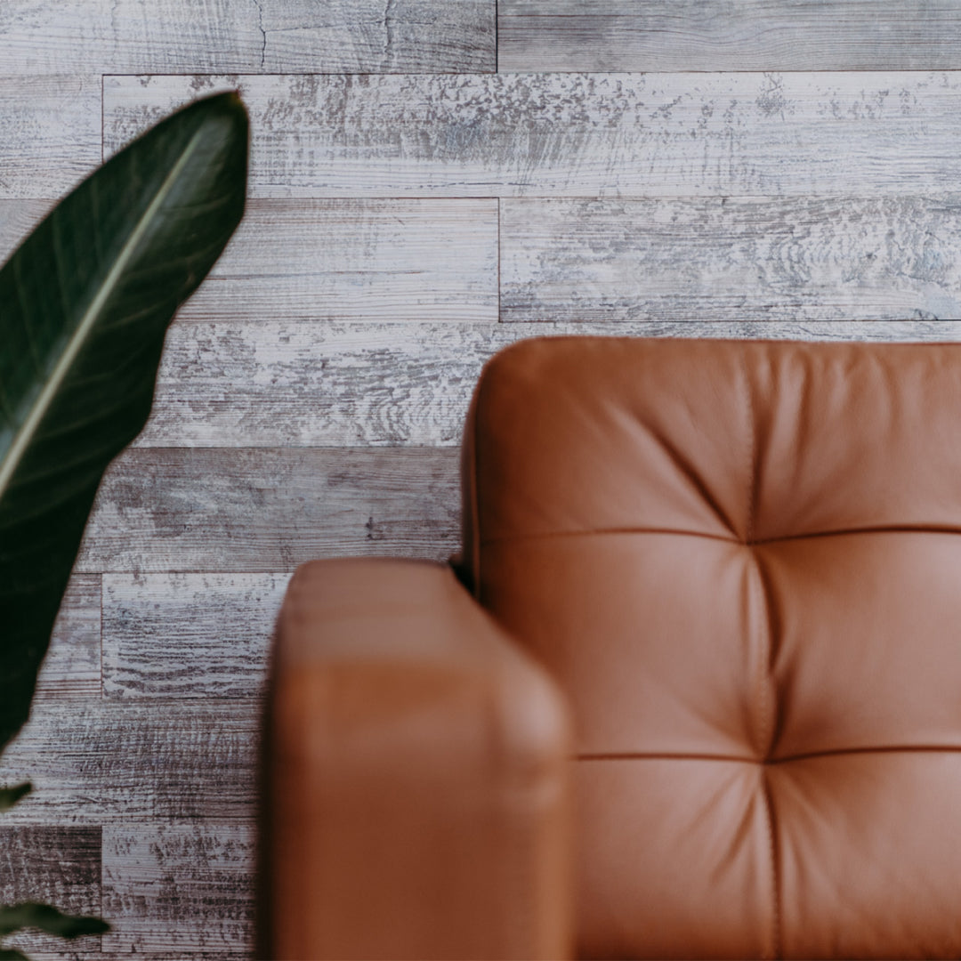 Ein braunes, gestepptes Leder-Sofa ist vor einer Wandverkleidung in grauer Vintage-Holzoptik platziert. Ein großes grünes Blatt einer Zimmerpflanze ragt links ins Bild und unterstreicht den modernen, wohnlichen Einrichtungsstil.