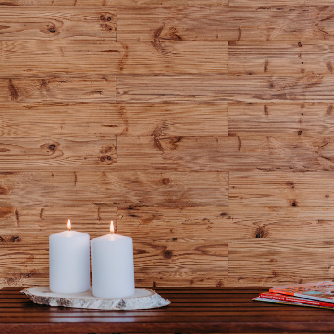 Zwei brennende Stumpenkerzen stehen auf einer rustikalen Baumscheibe, platziert auf einer dunklen Holzbank. Die Wand dahinter ist mit warmtönigen Holzpaneelen verkleidet, deren ausgeprägte Maserung eine natürliche und behagliche Raumstimmung erzeugt.