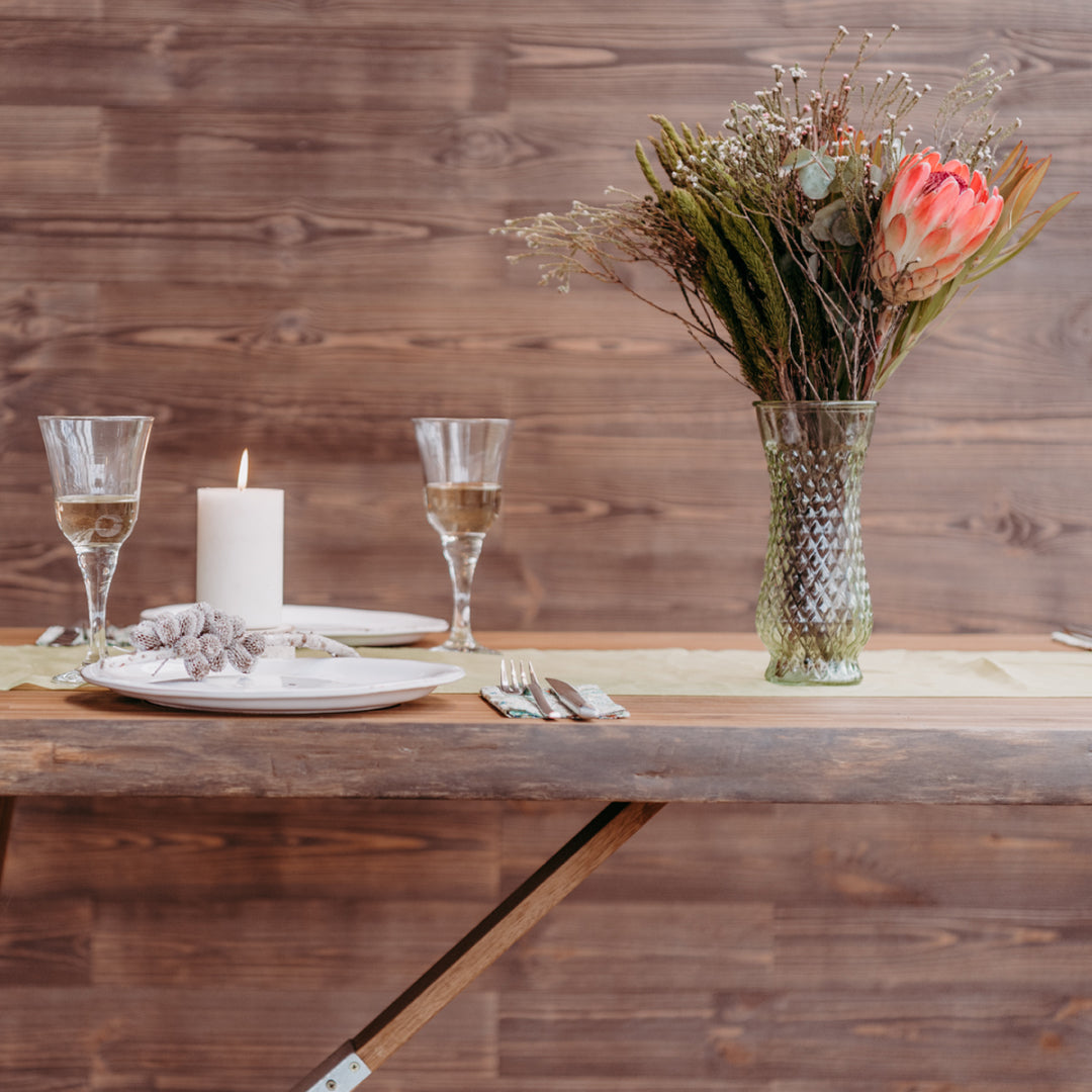 Ein massiver Echtholztisch ist festlich mit Weingläsern, einer Kerze und einer hohen Glasvase mit Trockenblumen gedeckt. Die horizontale Holzverkleidung im Hintergrund unterstreicht den warmen, rustikalen Charakter des Essbereichs.